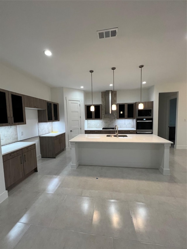 kitchen featuring tasteful backsplash, an island with sink, sink, dark brown cabinetry, and wall chimney range hood