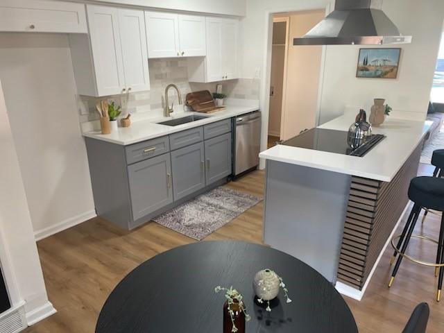 kitchen featuring a kitchen breakfast bar, stainless steel dishwasher, sink, wall chimney range hood, and gray cabinets