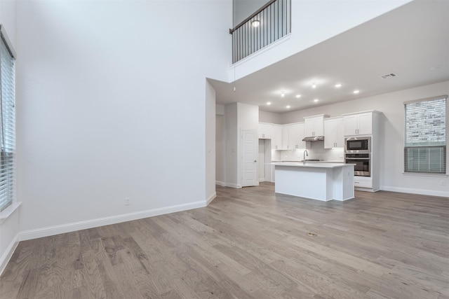 kitchen with light hardwood / wood-style flooring, an island with sink, a towering ceiling, stainless steel appliances, and white cabinets