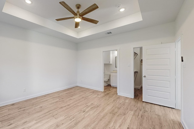 unfurnished bedroom with a tray ceiling, ensuite bathroom, ceiling fan, and light wood-type flooring