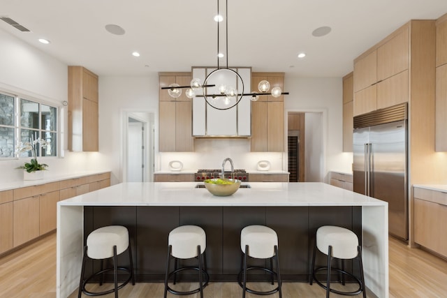 kitchen with stainless steel built in refrigerator, light brown cabinets, light hardwood / wood-style floors, and an island with sink