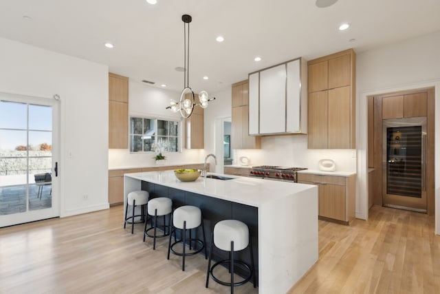 kitchen with a center island with sink, sink, hanging light fixtures, wine cooler, and light hardwood / wood-style flooring