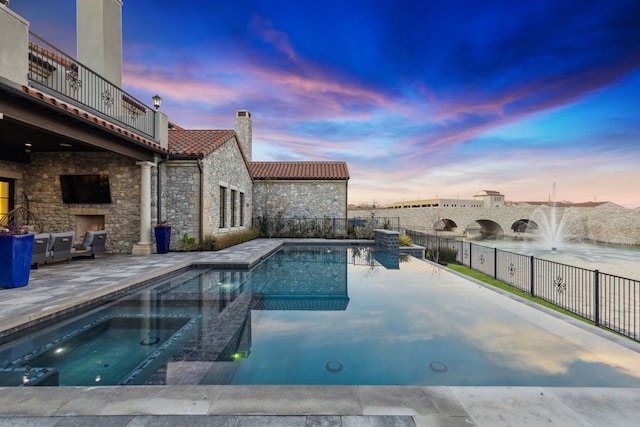 pool at dusk featuring an in ground hot tub, an outdoor stone fireplace, and a patio