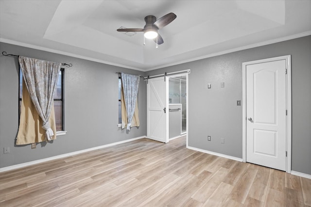 unfurnished bedroom featuring a raised ceiling, ceiling fan, a barn door, ornamental molding, and light hardwood / wood-style floors