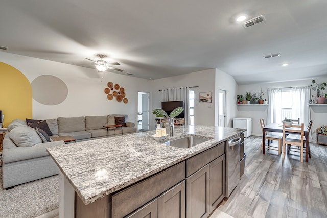 kitchen with ceiling fan, sink, light wood-type flooring, light stone counters, and a center island with sink