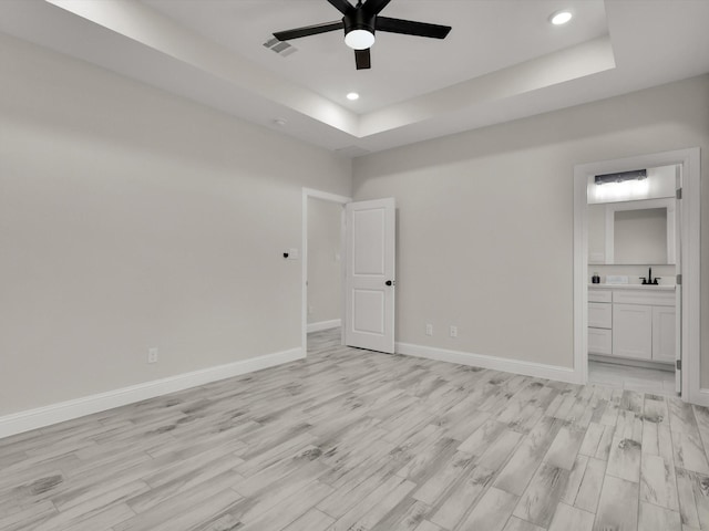 unfurnished bedroom featuring sink, ensuite bath, ceiling fan, a tray ceiling, and light hardwood / wood-style floors