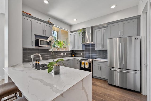 kitchen with kitchen peninsula, light stone counters, wall chimney exhaust hood, stainless steel appliances, and gray cabinets