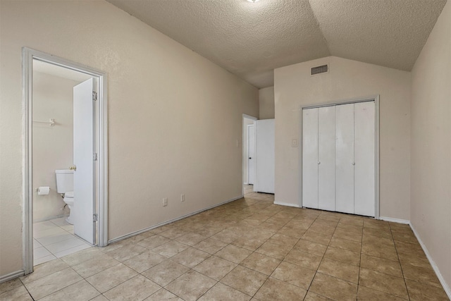 unfurnished bedroom featuring ensuite bathroom, a textured ceiling, a closet, lofted ceiling, and light tile patterned flooring