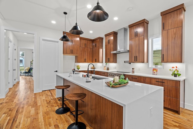 kitchen with wall chimney range hood, sink, light hardwood / wood-style flooring, an island with sink, and decorative light fixtures