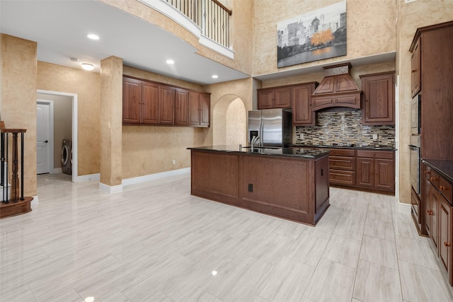 kitchen with a high ceiling, washer / dryer, a center island with sink, custom exhaust hood, and appliances with stainless steel finishes