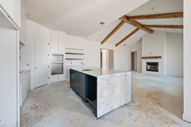 kitchen with vaulted ceiling with beams, light stone counters, white cabinets, and an island with sink