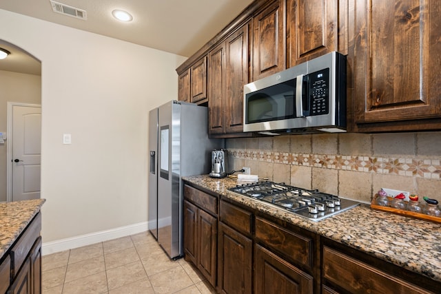 kitchen with stainless steel appliances, dark brown cabinetry, light tile patterned flooring, decorative backsplash, and dark stone counters