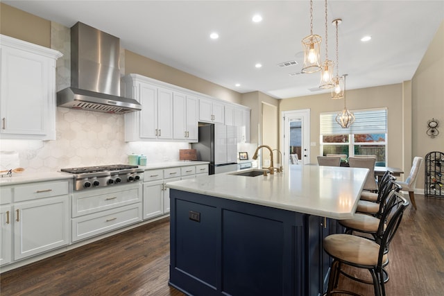 kitchen with sink, wall chimney exhaust hood, stainless steel appliances, a kitchen island with sink, and white cabinets