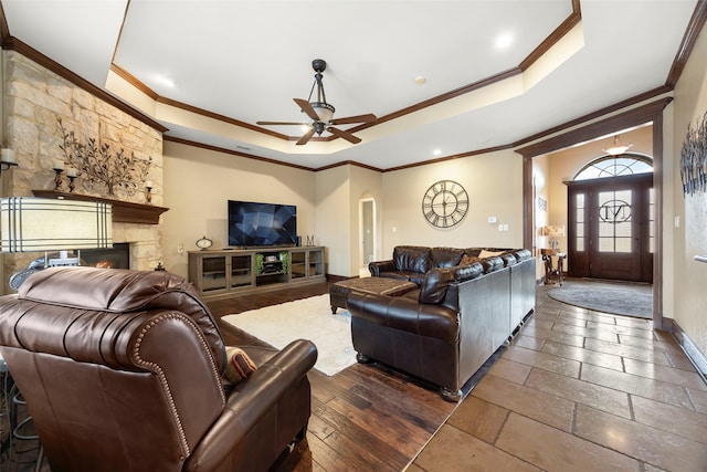 living room featuring a tray ceiling, a stone fireplace, ceiling fan, and ornamental molding