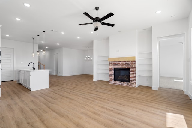 unfurnished living room featuring a brick fireplace, ceiling fan with notable chandelier, sink, built in features, and light hardwood / wood-style flooring