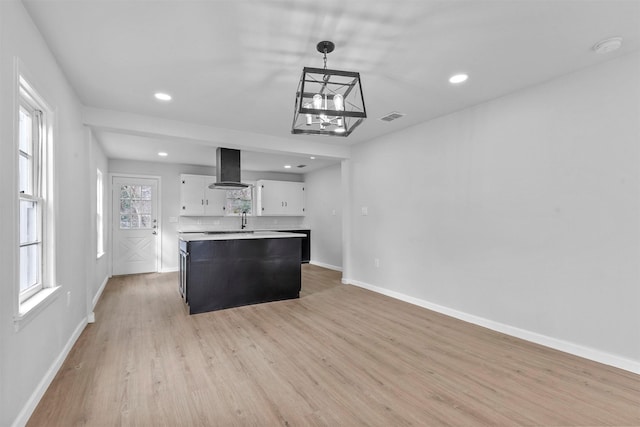 kitchen featuring a center island, wall chimney range hood, hanging light fixtures, light wood-type flooring, and white cabinetry