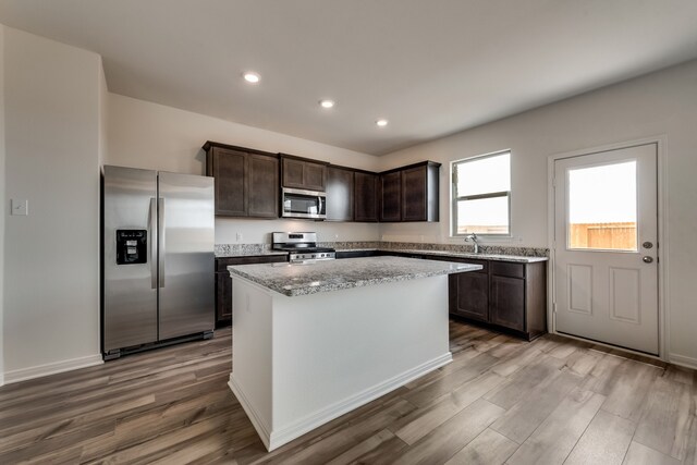 kitchen with light stone countertops, dark brown cabinetry, stainless steel appliances, hardwood / wood-style flooring, and a kitchen island