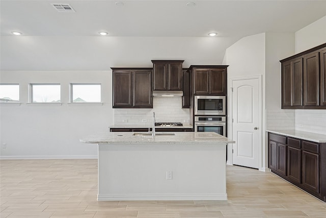 kitchen with dark brown cabinets, light stone countertops, an island with sink, and appliances with stainless steel finishes