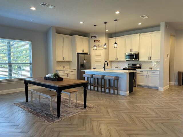 kitchen featuring stainless steel appliances, light parquet floors, decorative light fixtures, white cabinets, and an island with sink