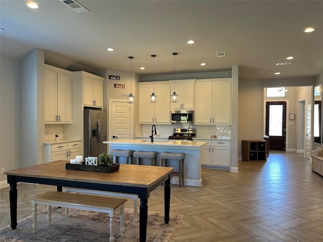 kitchen with light parquet floors, decorative light fixtures, a center island with sink, white cabinets, and appliances with stainless steel finishes