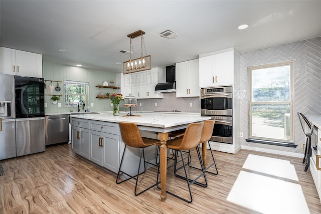 kitchen featuring decorative light fixtures, a center island with sink, wall chimney range hood, stainless steel appliances, and white cabinets