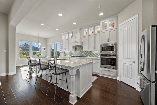 kitchen featuring stainless steel appliances, premium range hood, an island with sink, decorative light fixtures, and white cabinets