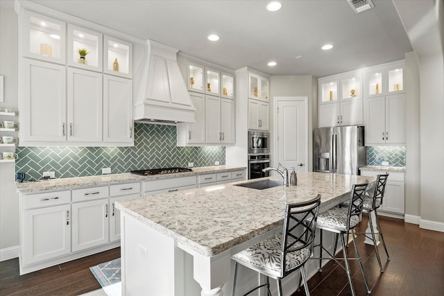 kitchen featuring white cabinets, custom range hood, an island with sink, and appliances with stainless steel finishes