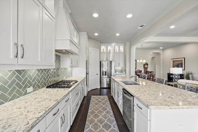 kitchen featuring custom exhaust hood, white cabinets, a center island with sink, sink, and appliances with stainless steel finishes