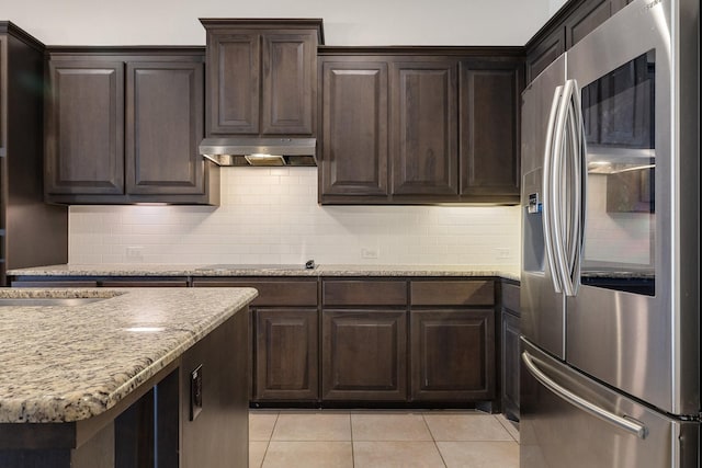 kitchen featuring light tile patterned floors, stainless steel fridge, dark brown cabinets, light stone counters, and decorative backsplash