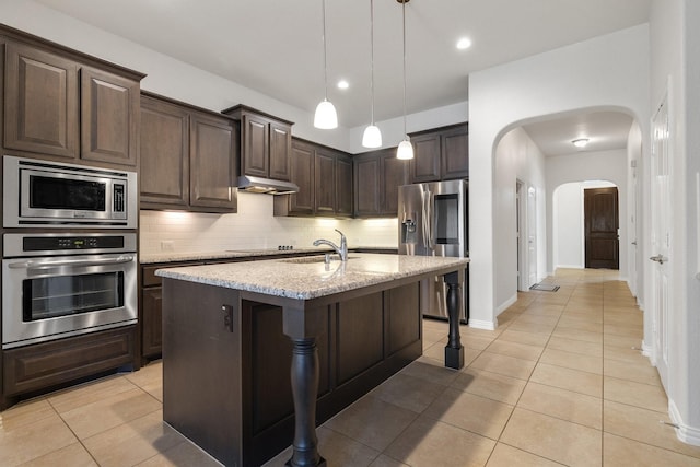 kitchen featuring appliances with stainless steel finishes, decorative light fixtures, decorative backsplash, light stone counters, and a center island with sink