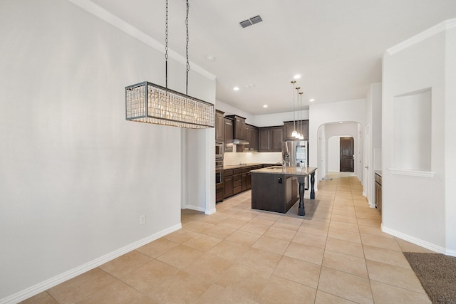 kitchen featuring a breakfast bar, decorative light fixtures, crown molding, dark brown cabinets, and a center island with sink