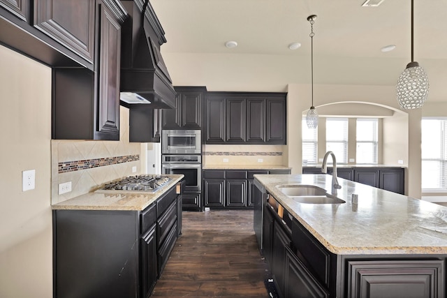 kitchen featuring sink, custom exhaust hood, hanging light fixtures, appliances with stainless steel finishes, and an island with sink