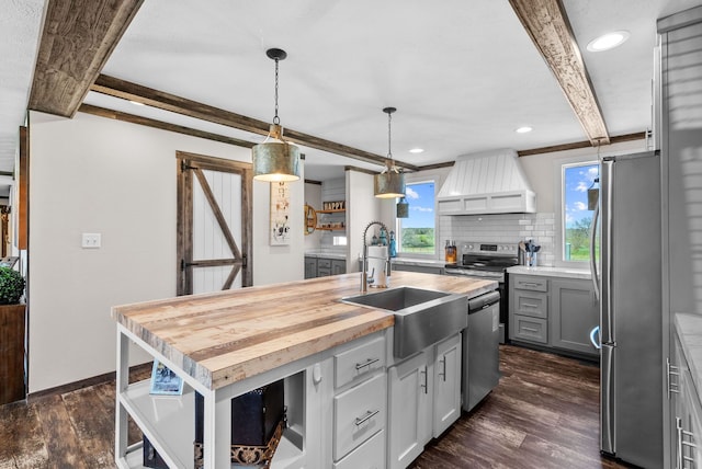 kitchen featuring a sink, wooden counters, appliances with stainless steel finishes, custom exhaust hood, and a center island with sink