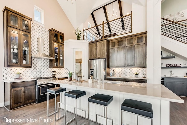 kitchen featuring high end appliances, wall chimney range hood, a center island with sink, high vaulted ceiling, and hardwood / wood-style floors