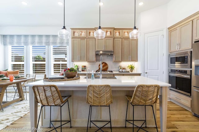 kitchen with light brown cabinets, light hardwood / wood-style flooring, backsplash, an island with sink, and appliances with stainless steel finishes