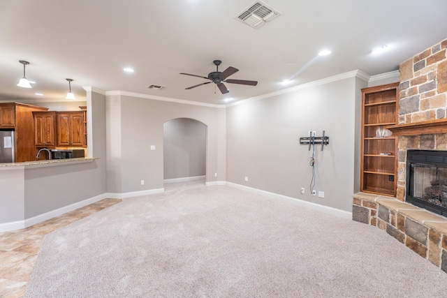 unfurnished living room with ceiling fan, sink, a stone fireplace, light colored carpet, and ornamental molding