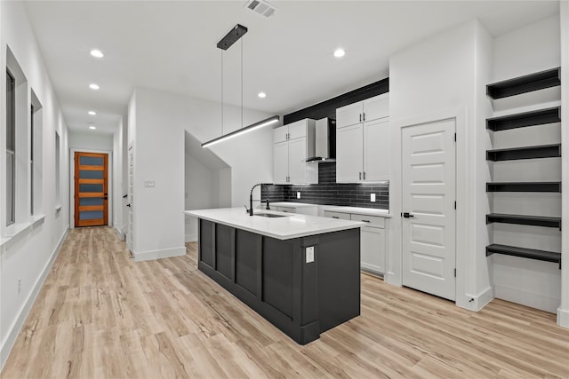 kitchen with pendant lighting, a center island with sink, wall chimney exhaust hood, light hardwood / wood-style floors, and white cabinetry