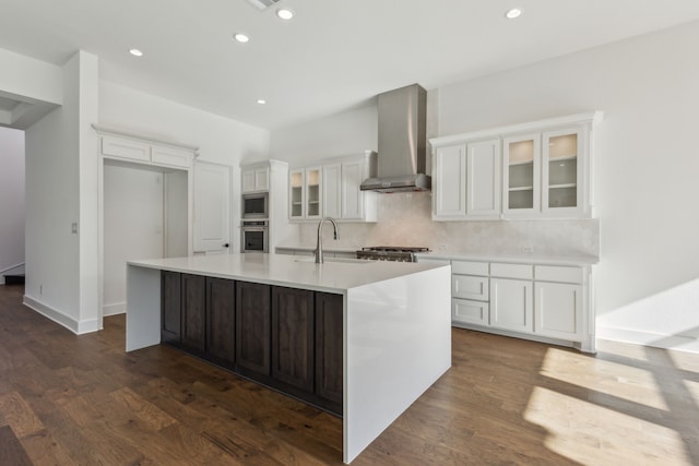 kitchen featuring stainless steel appliances, wall chimney range hood, tasteful backsplash, an island with sink, and white cabinets