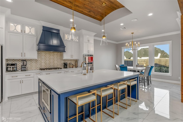 kitchen featuring premium range hood, tasteful backsplash, white cabinetry, crown molding, and a center island with sink