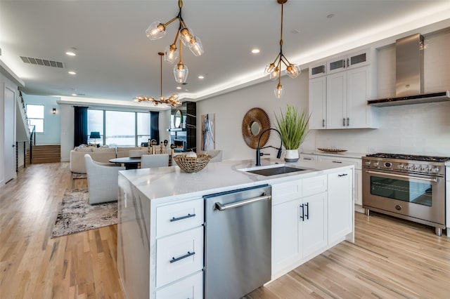 kitchen with white cabinets, wall chimney range hood, sink, an island with sink, and appliances with stainless steel finishes