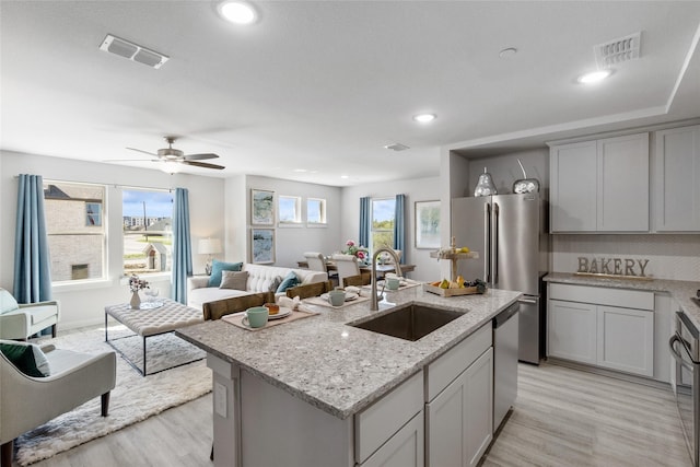 kitchen with ceiling fan, sink, light stone counters, a center island with sink, and appliances with stainless steel finishes