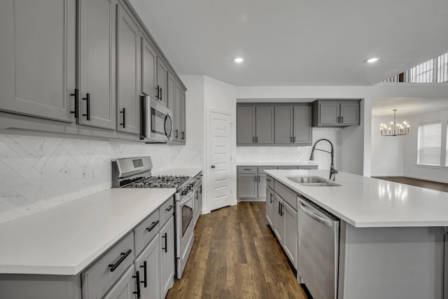 kitchen with gray cabinetry, sink, stainless steel appliances, an inviting chandelier, and a center island with sink