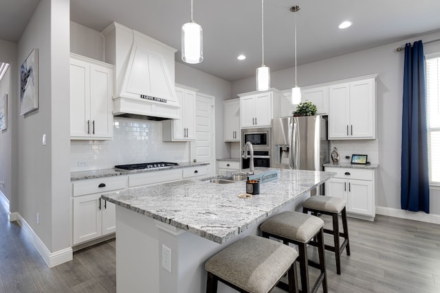 kitchen featuring appliances with stainless steel finishes, custom range hood, white cabinets, an island with sink, and decorative light fixtures