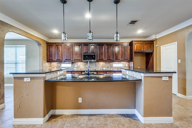 kitchen with tasteful backsplash, a center island with sink, hanging light fixtures, and dark stone counters