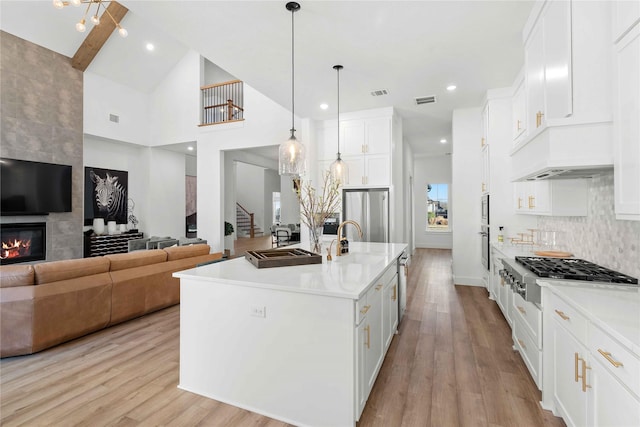 kitchen featuring white cabinetry, decorative light fixtures, a large fireplace, appliances with stainless steel finishes, and an island with sink