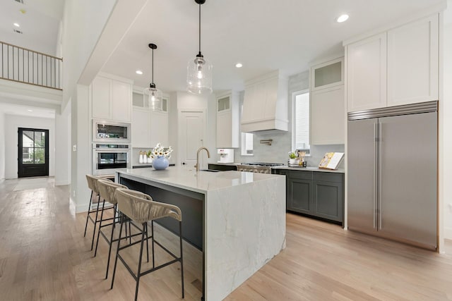 kitchen with white cabinets, built in appliances, custom range hood, and a center island with sink