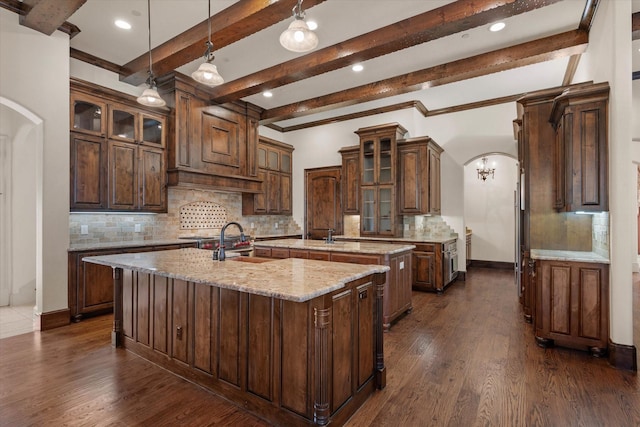 kitchen with light stone counters, beam ceiling, a center island with sink, dark hardwood / wood-style floors, and hanging light fixtures