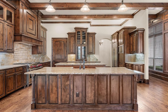 kitchen featuring beamed ceiling, stainless steel stove, hanging light fixtures, and an island with sink