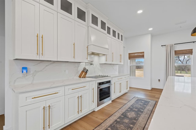 kitchen featuring tasteful backsplash, light stone counters, custom exhaust hood, white cabinets, and oven