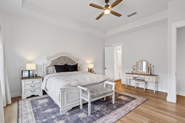 bedroom featuring ceiling fan, light wood-type flooring, ensuite bathroom, and a tray ceiling